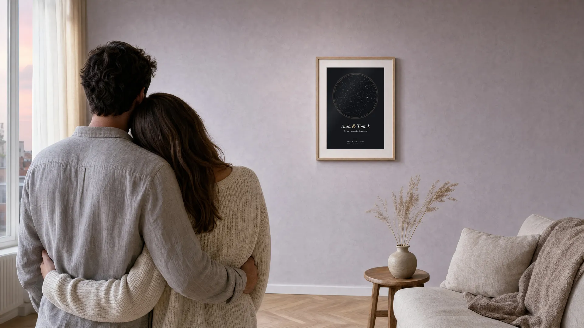 Para przytulona o świcie w salonie patrzy na granatowy plakat z mapą nieba w dębowej ramie